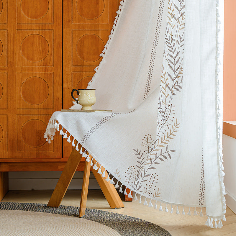 White embroidered curtains with tassels draped over a wooden chair against a wooden cabinet.