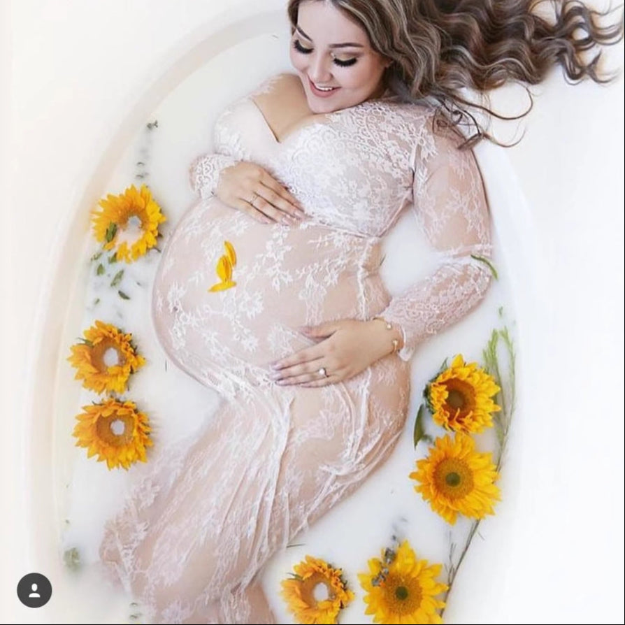 Pregnant woman in a white lace dress surrounded by sunflowers in a bathtub.