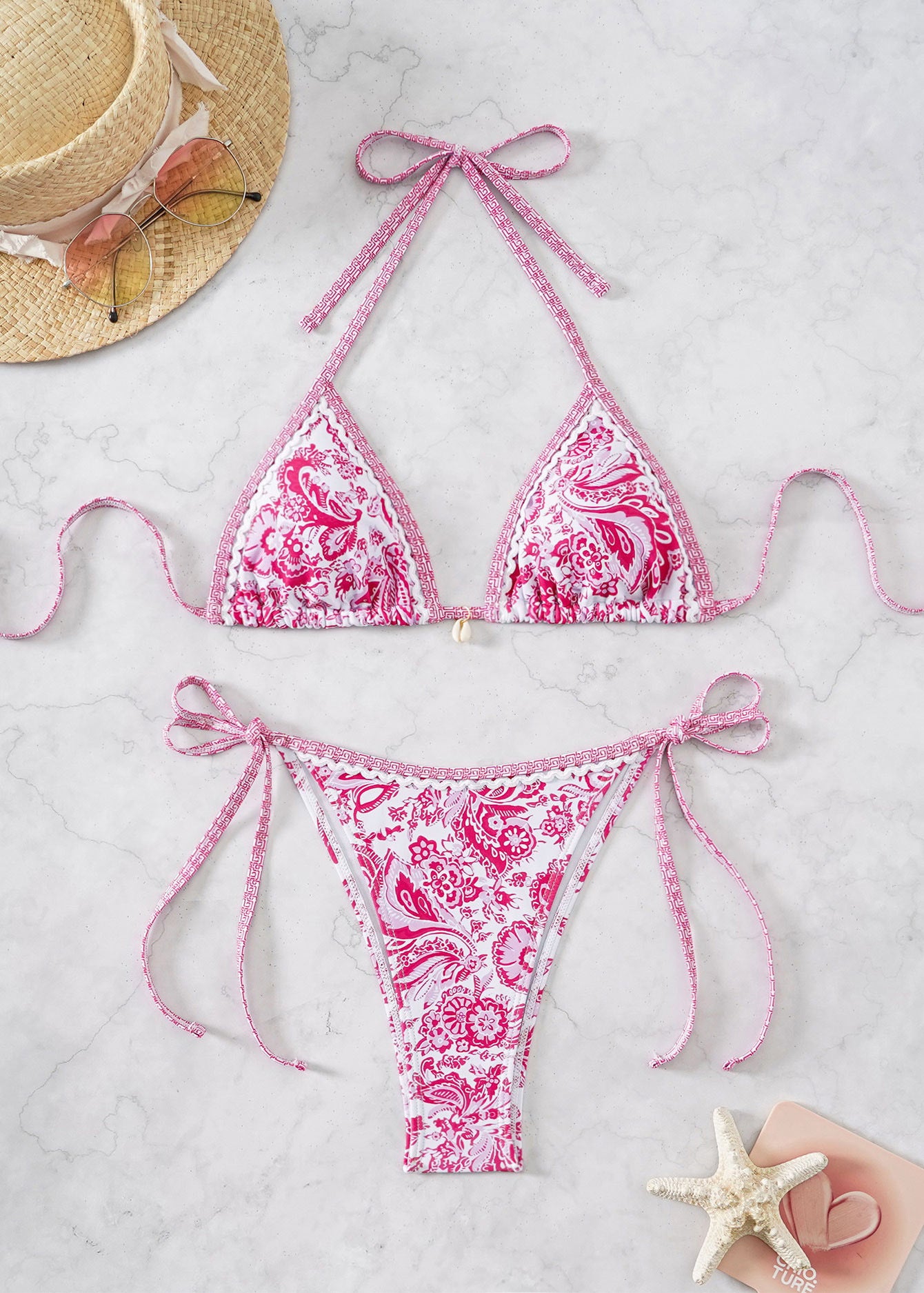 Pink patterned bikini set on a light gray surface with a straw hat and starfish.