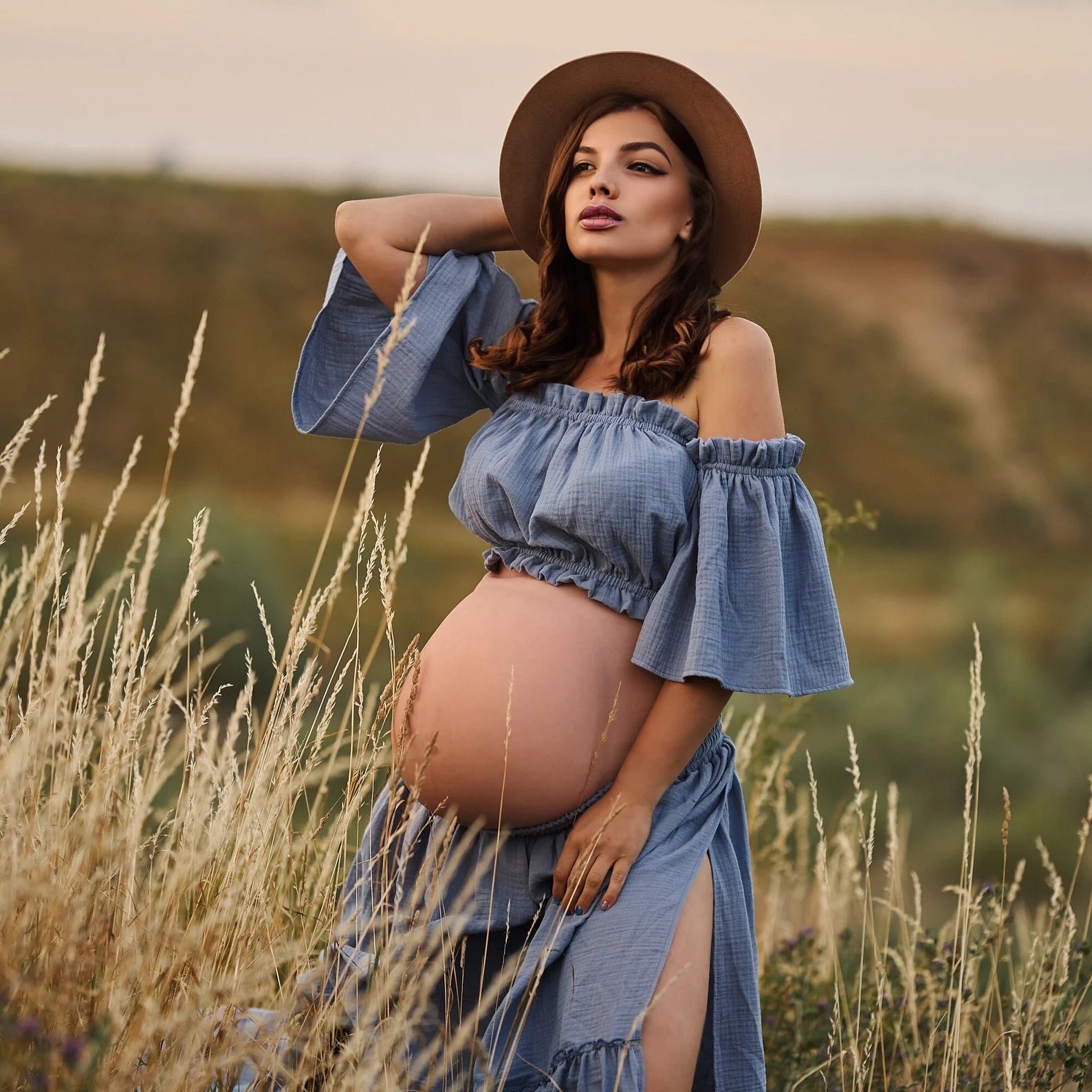 Pregnant woman in a blue dress and hat standing in a field.