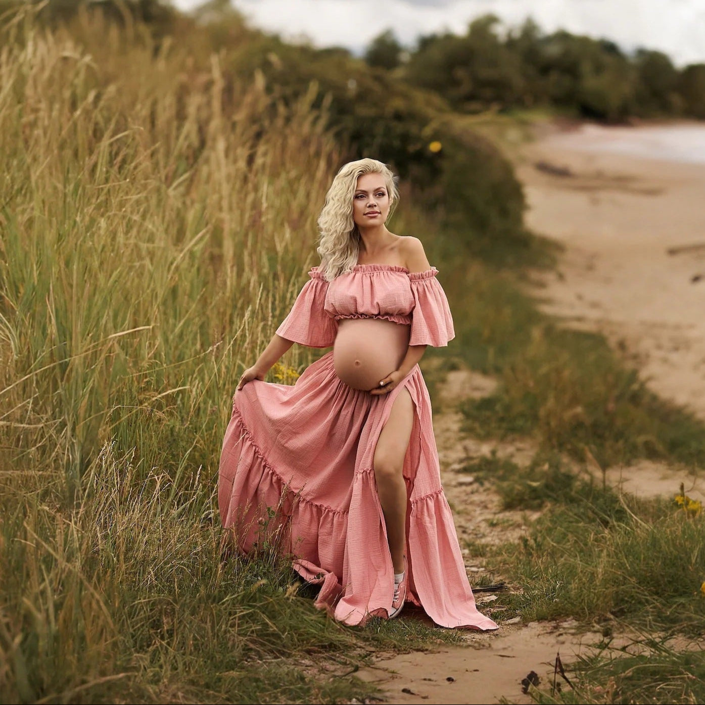 Pregnant woman in a pink dress standing in a grassy field near a beach.