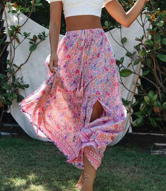 Woman wearing a white top and colorful skirt standing outdoors with greenery in the background