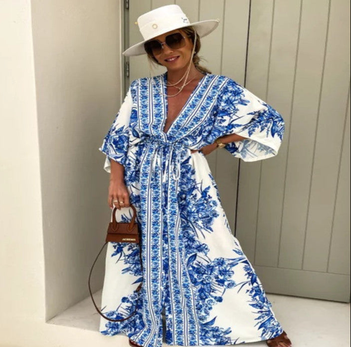 Woman wearing a blue and white floral dress with a white hat and sunglasses, standing against a light-colored wall.