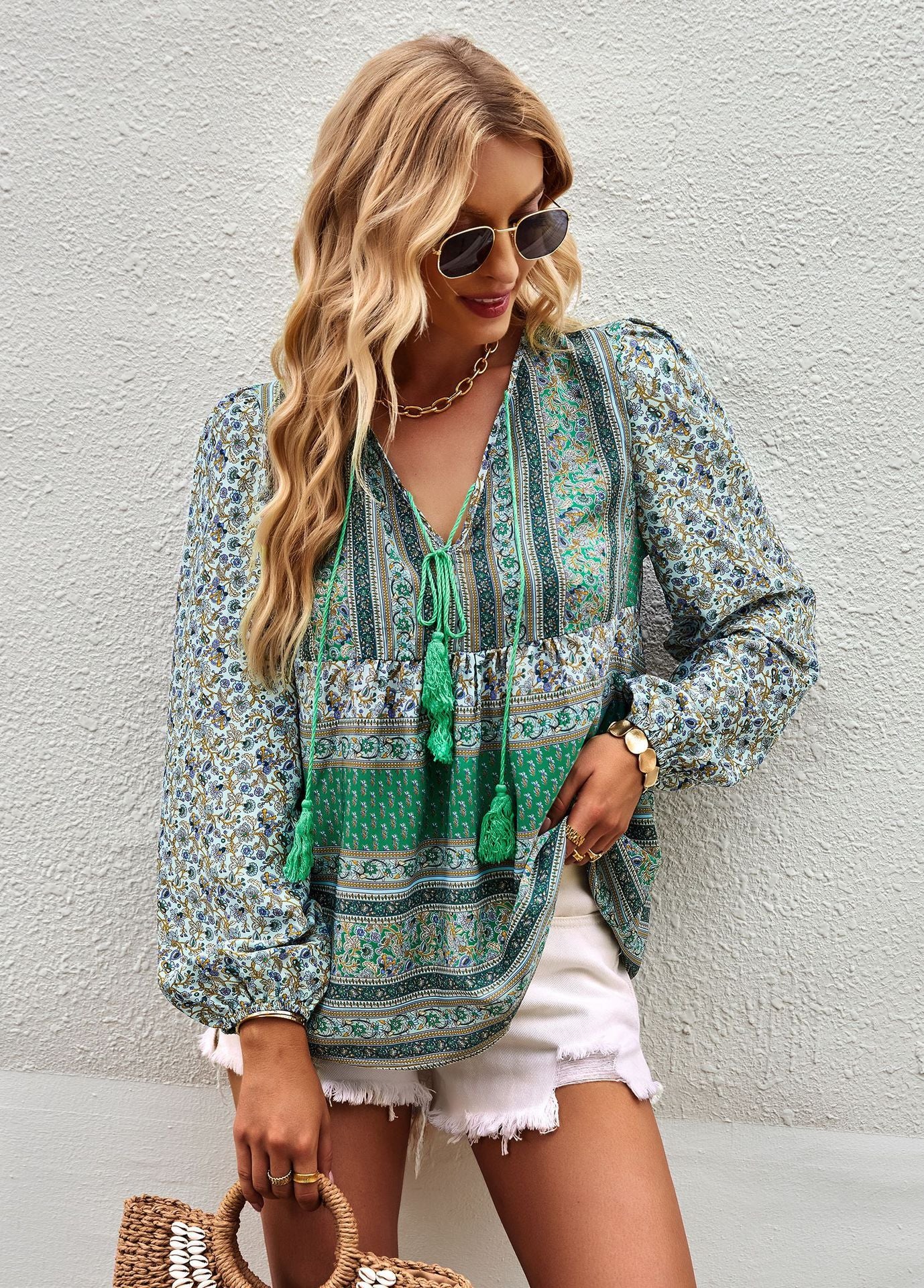 Woman wearing a green patterned blouse with white shorts against a light gray wall.