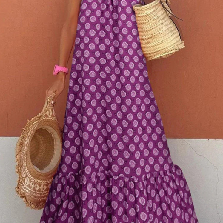 Person wearing a purple dress with white patterns, holding a woven bag against a peach-colored wall.