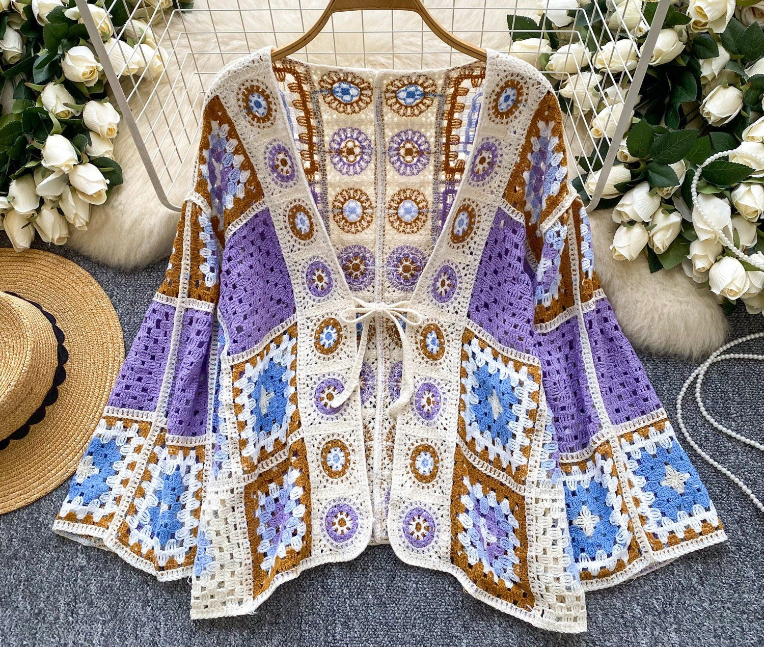 Crocheted cardigan with blue, brown, and white pattern on a hanger with flowers and a hat in the background.