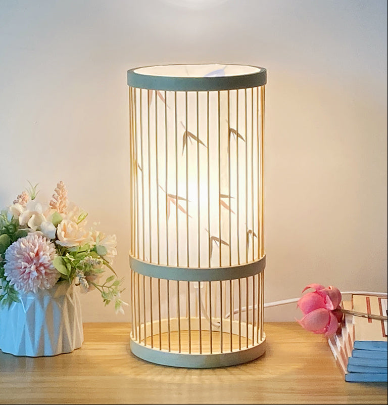 Cylindrical table lamp with bamboo design on a wooden surface, next to flowers and books.