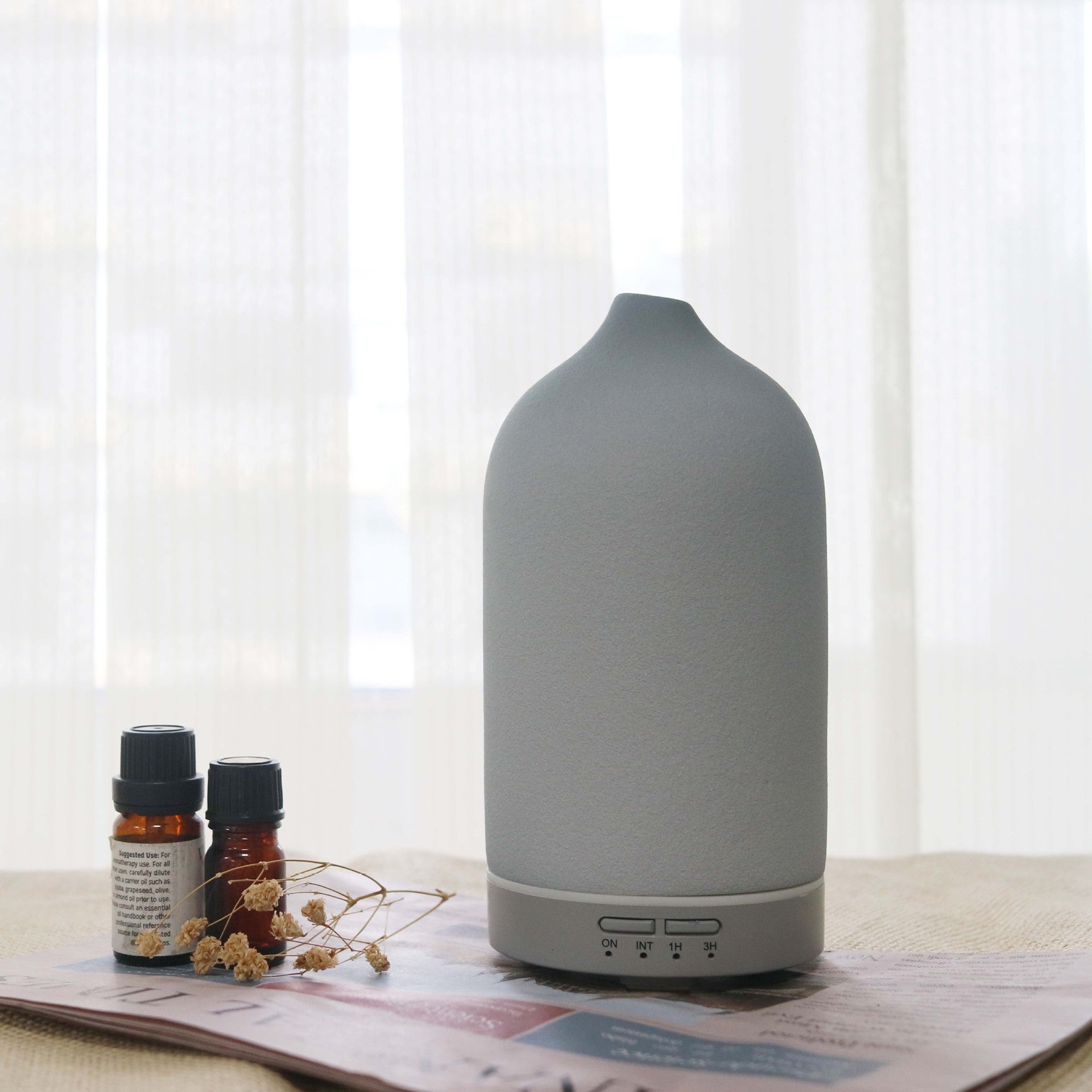 Gray diffuser on a table with essential oil bottles and dried flowers, against a white curtain background.