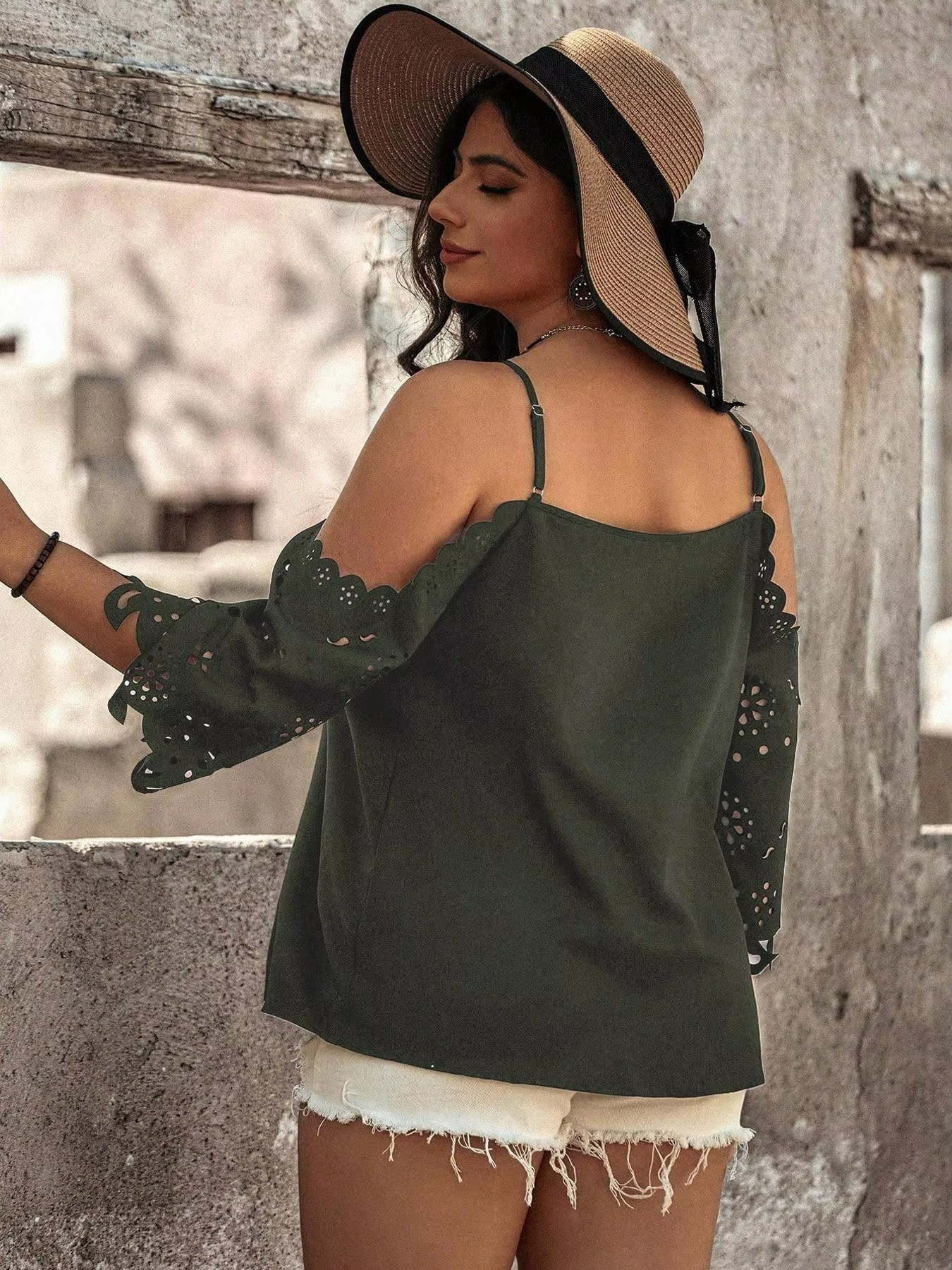 Woman wearing a green top with embellished sleeves and a wide-brimmed hat against a rustic wall.
