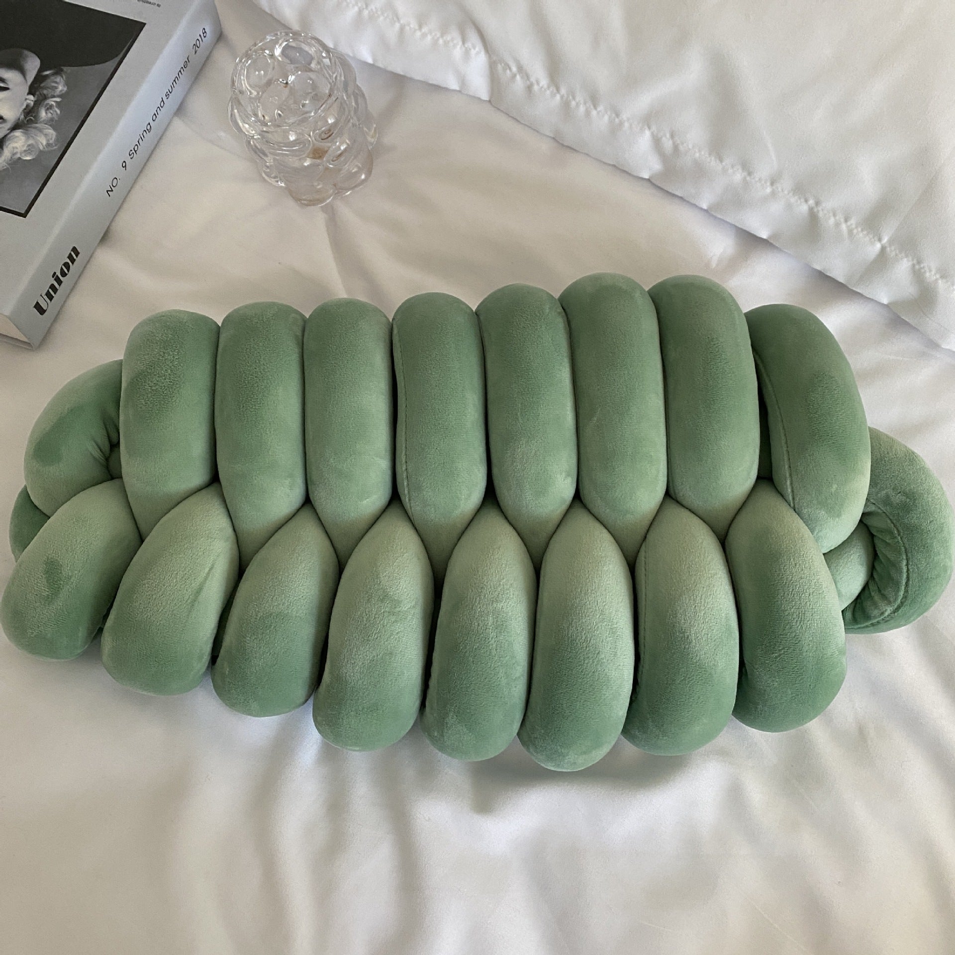 Green textured pillow on a white surface with a book and glass object in the background