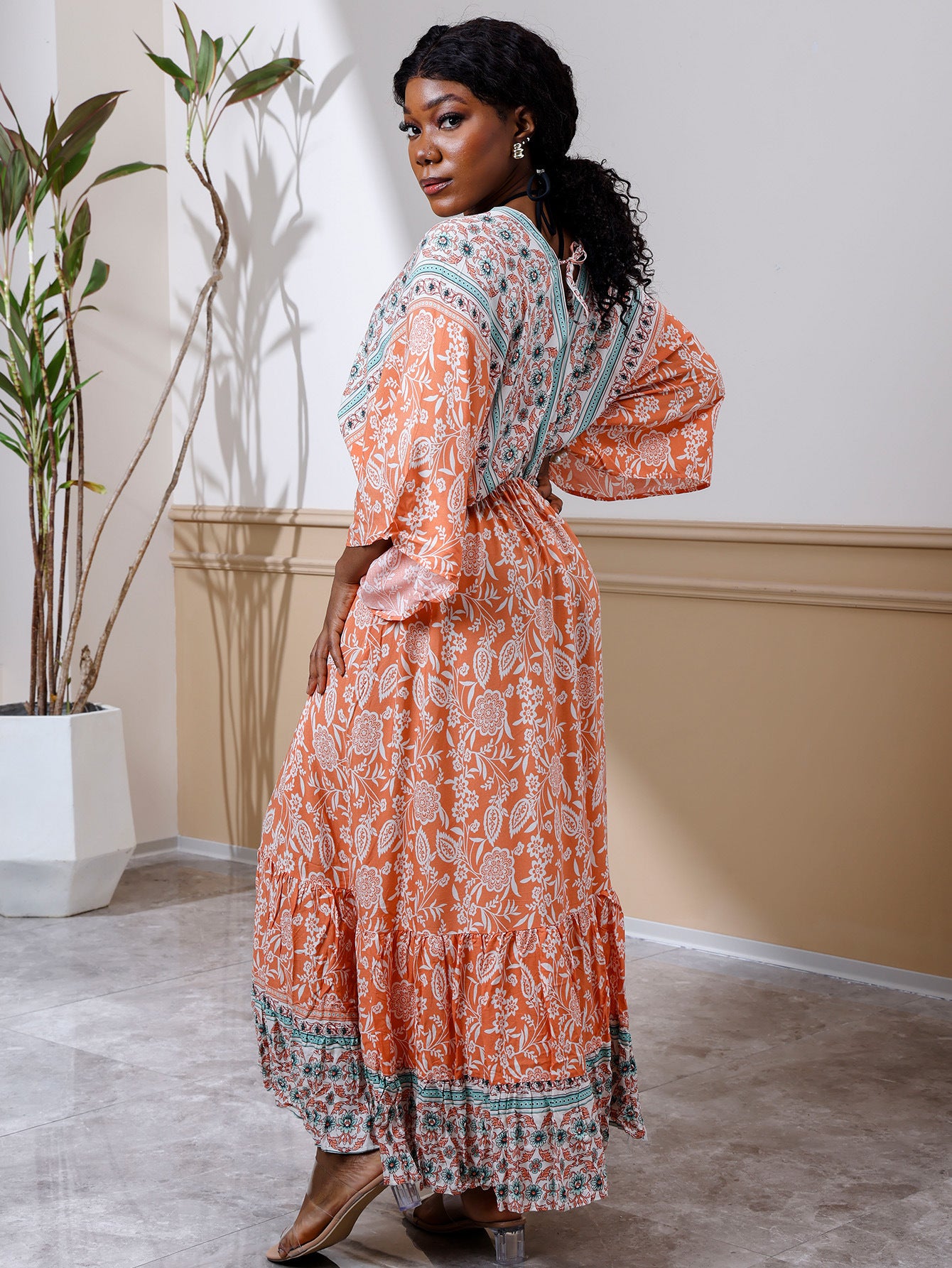 Woman in an orange patterned dress standing in a room with a plant and wall.