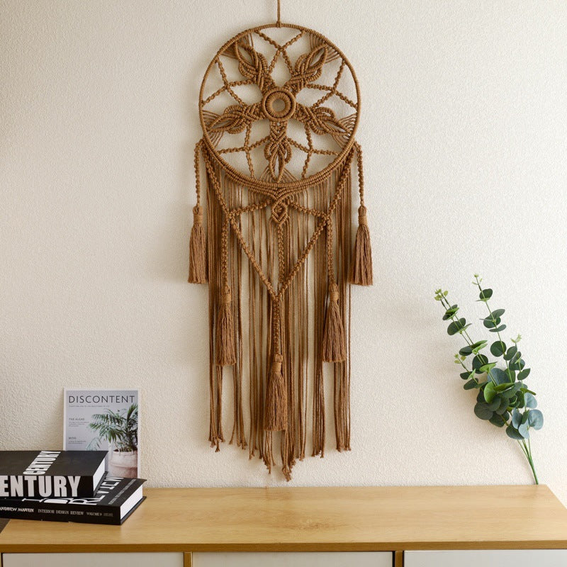 Brown dreamcatcher hanging on a white wall above a wooden surface with books and a plant.