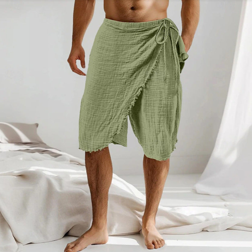 A man wearing light sage green gauze wrap shorts with a drawstring waist, standing barefoot in a bright, minimalist bedroom setting.