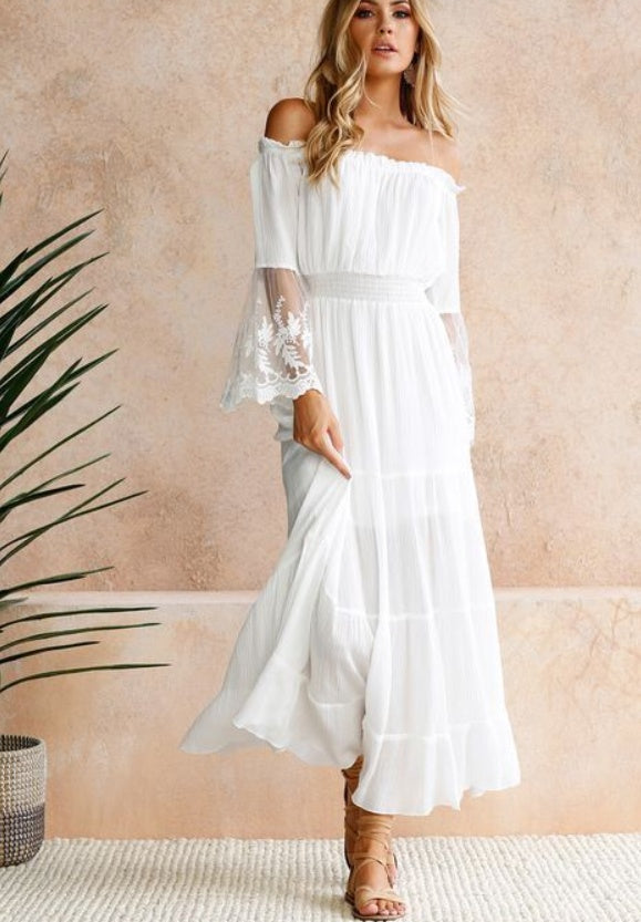 Woman wearing a white off-shoulder dress with lace sleeves against a beige wall.