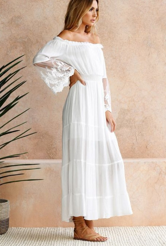 Woman wearing a white off-shoulder dress with sheer sleeves against a beige wall.