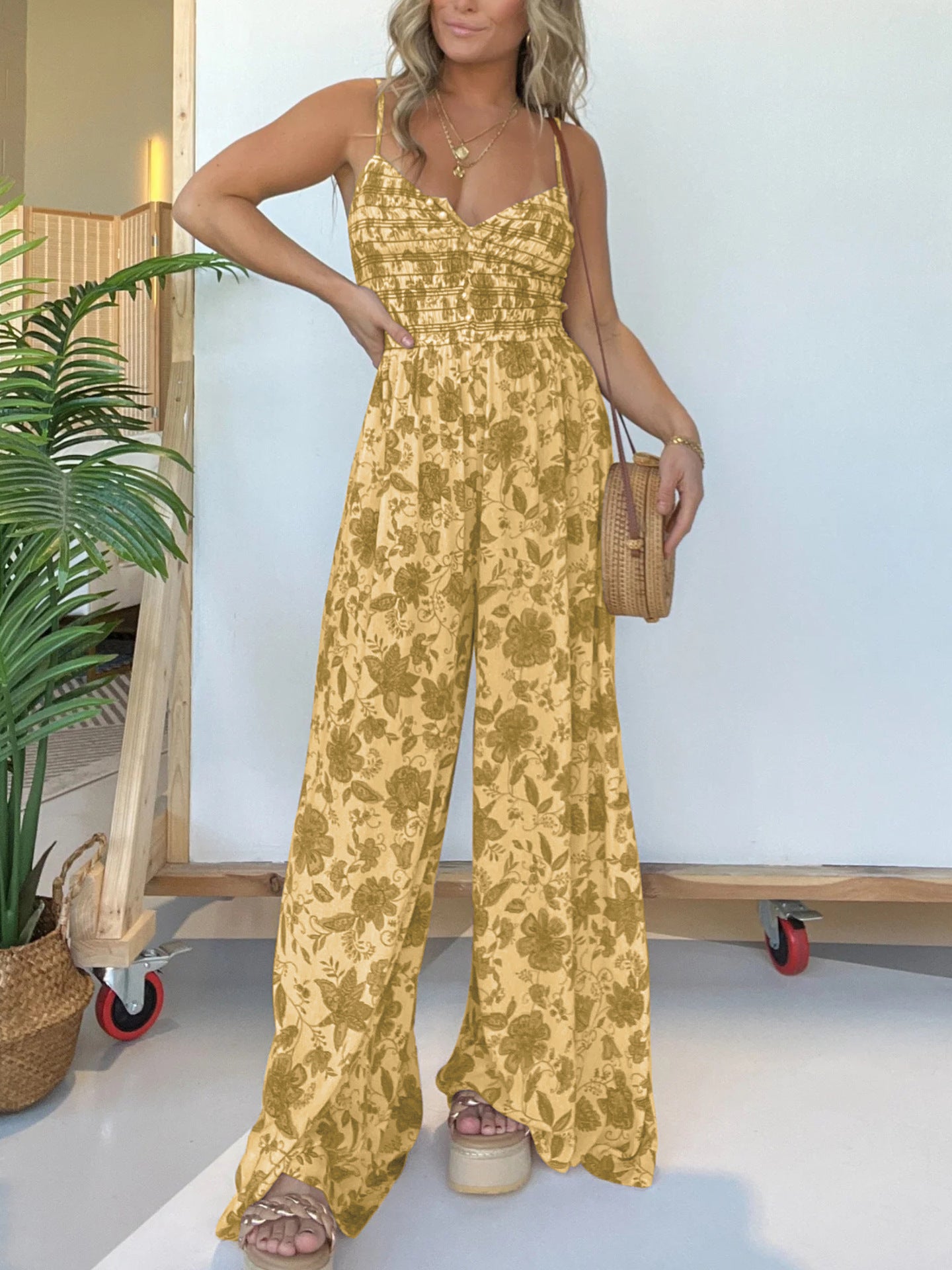 Woman wearing a yellow floral jumpsuit standing indoors with a plant and wooden cart in the background.