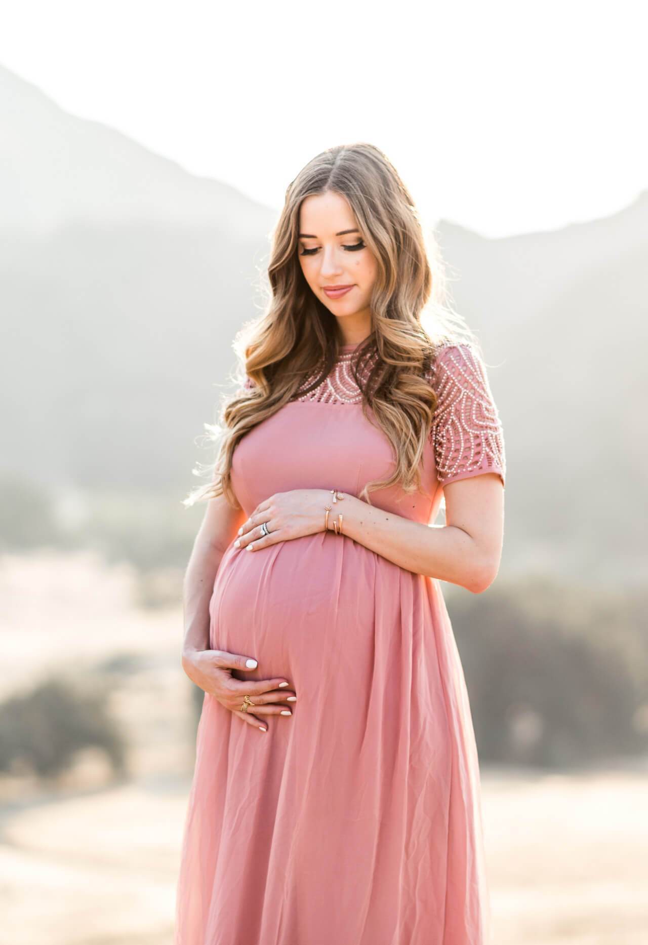 Pregnant woman in a pink dress standing outdoors with mountains in the background