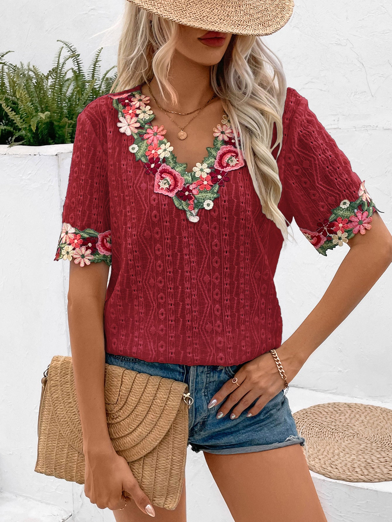 Woman wearing a red embroidered top with floral details, denim shorts, and a straw hat outdoors.