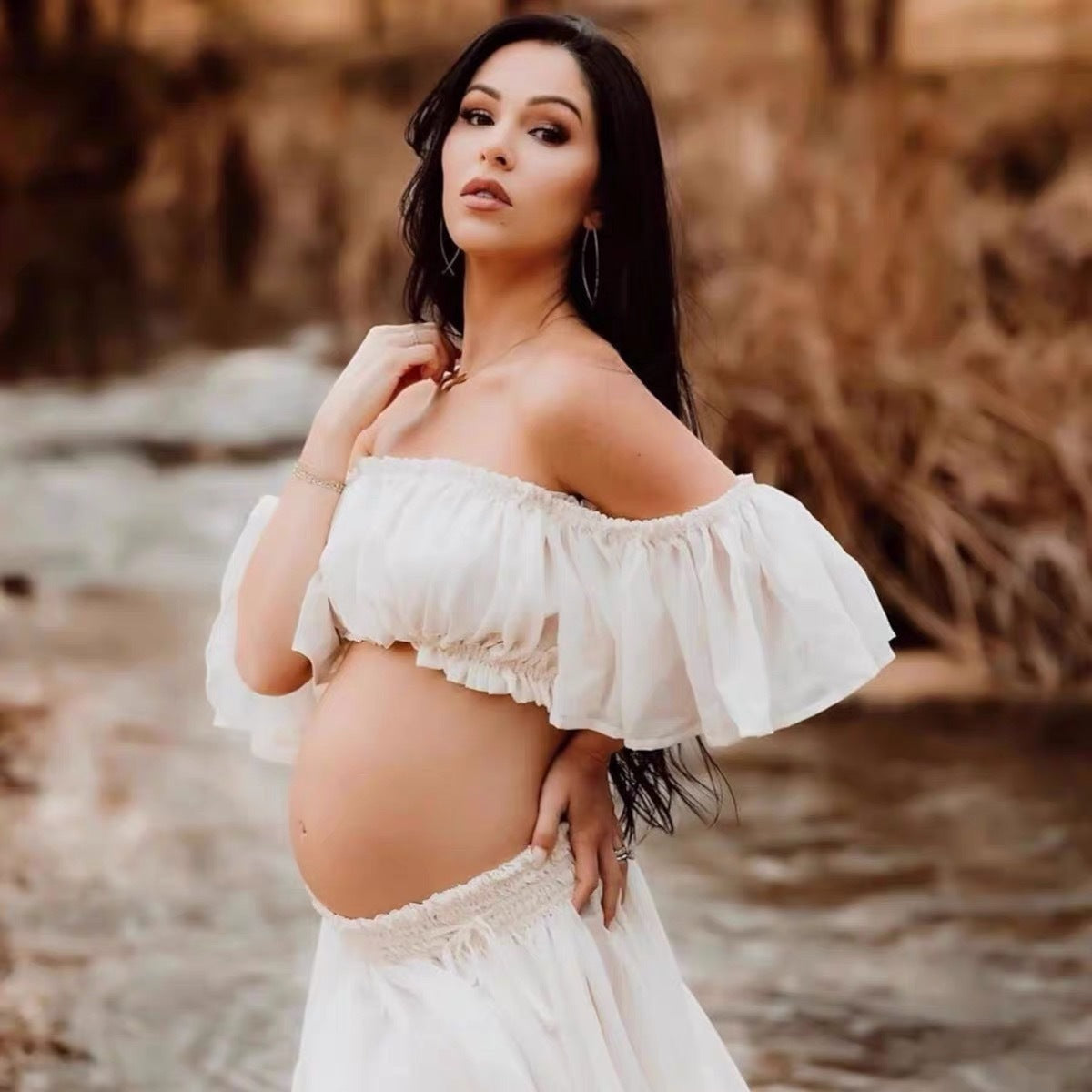 Pregnant woman in a white off-shoulder top and skirt standing by a body of water.