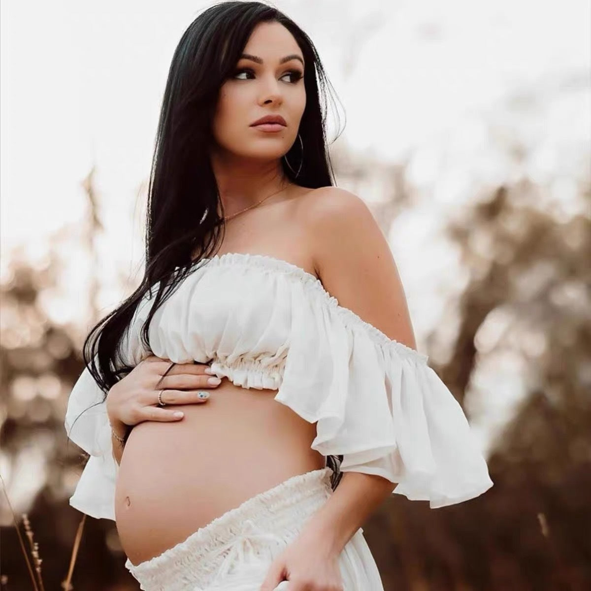 Pregnant woman in a white off-shoulder top and skirt standing outdoors with a blurred natural background