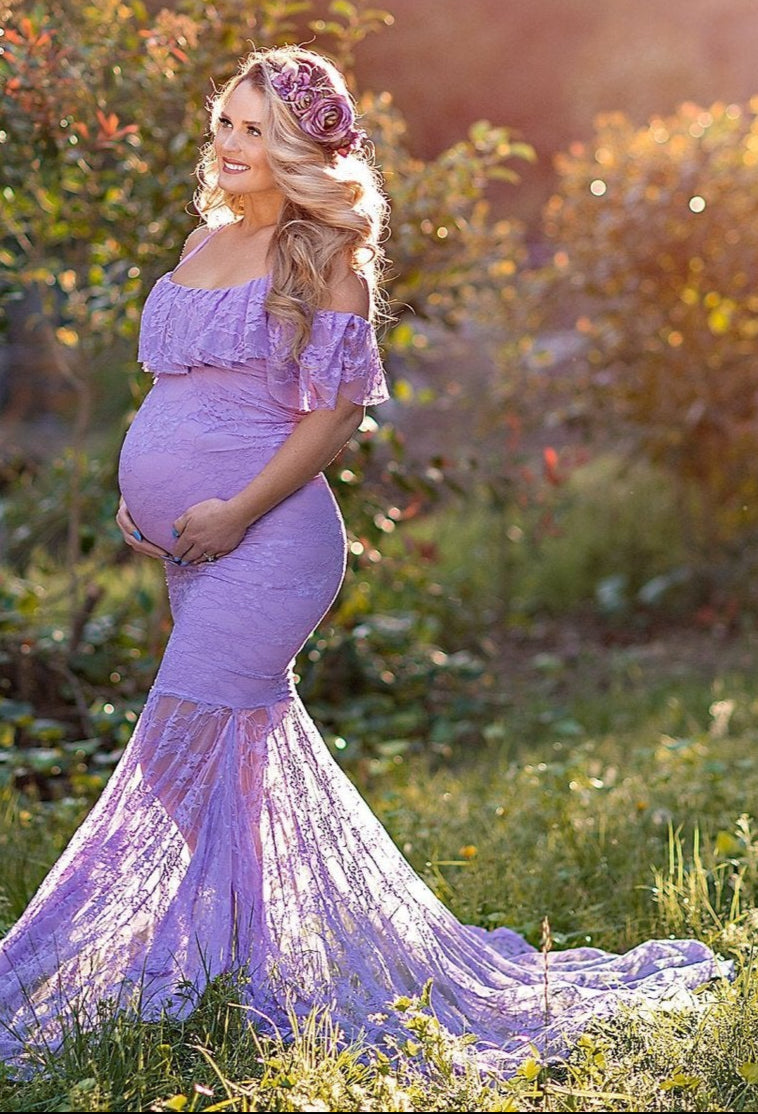 Pregnant woman in a purple gown standing in a garden with sunlight filtering through the trees.