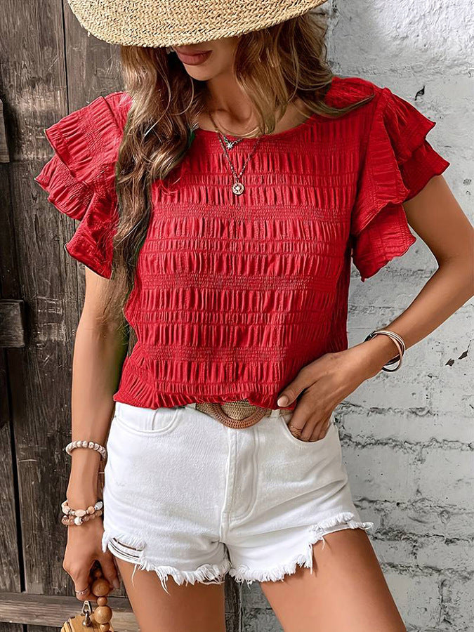 Woman wearing a red ruffled top and white shorts against a rustic wooden and stone wall.