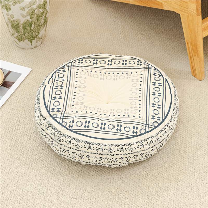 Round patterned cushion on a beige carpet with a wooden chair and vase in the background.