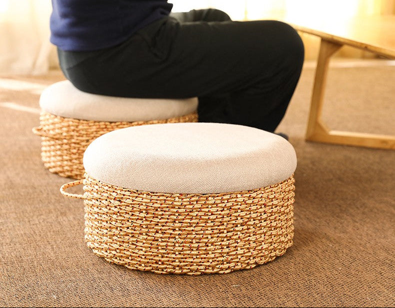 Person sitting on a woven ottoman with a cushion in a home setting.