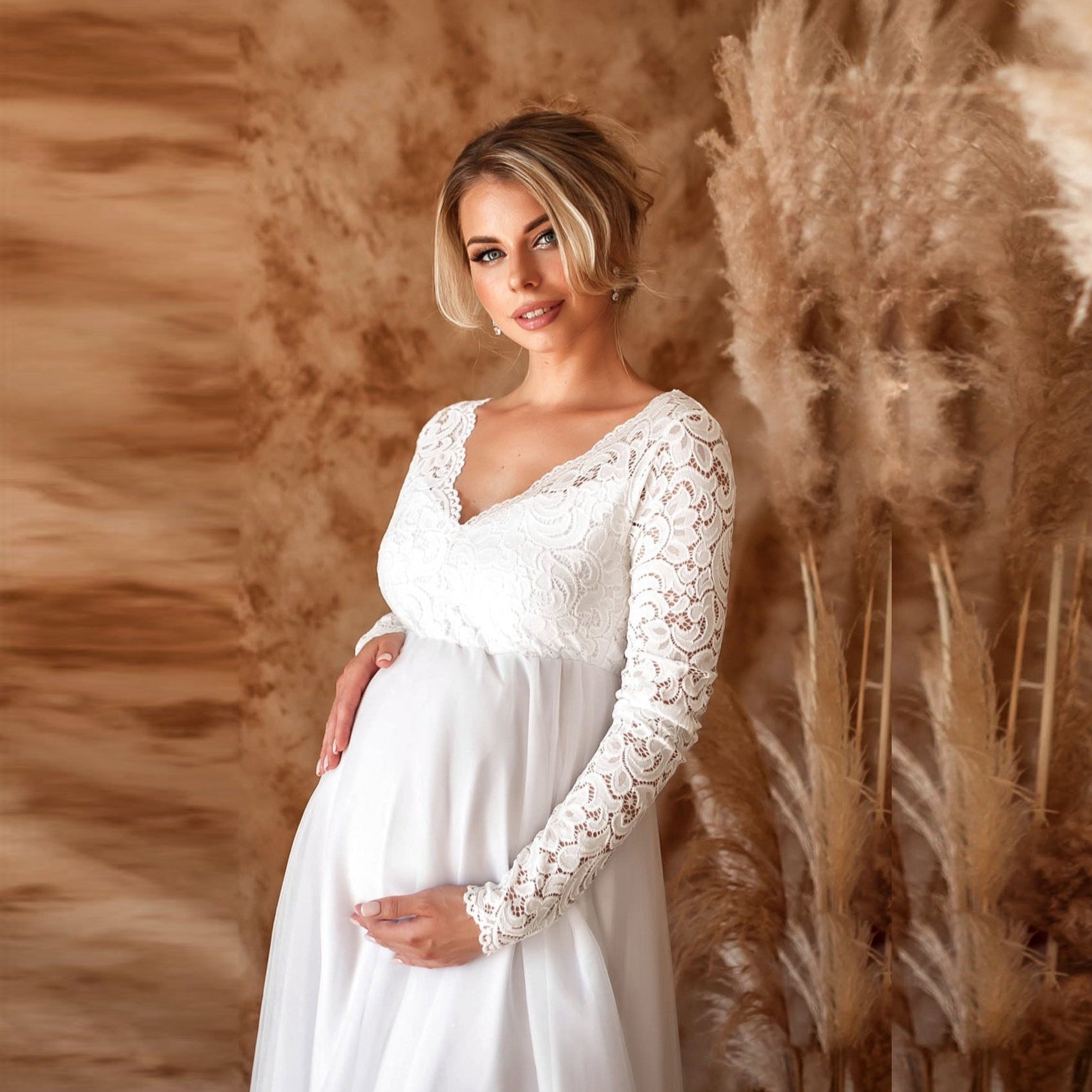 Pregnant woman in a white lace dress standing against a warm-toned background with pampas grass.