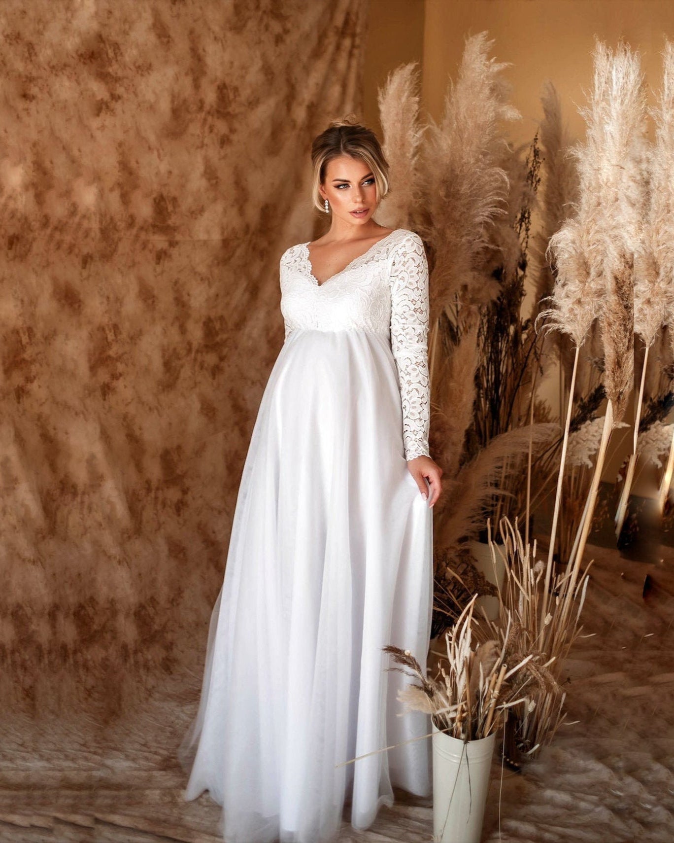 Woman in a white wedding dress standing among pampas grass against a textured wall.