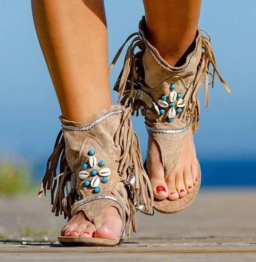 Beige suede sandals with fringe and blue embellishments on a blurred natural background
