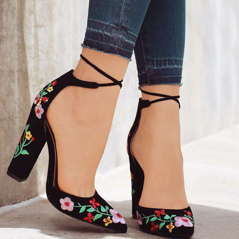 Black high-heeled shoes with floral embroidery worn with blue jeans on a neutral background