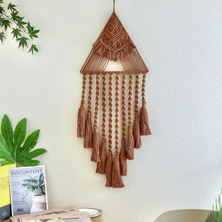 Brown macrame wall hanging on a white wall with plants and a magazine below.