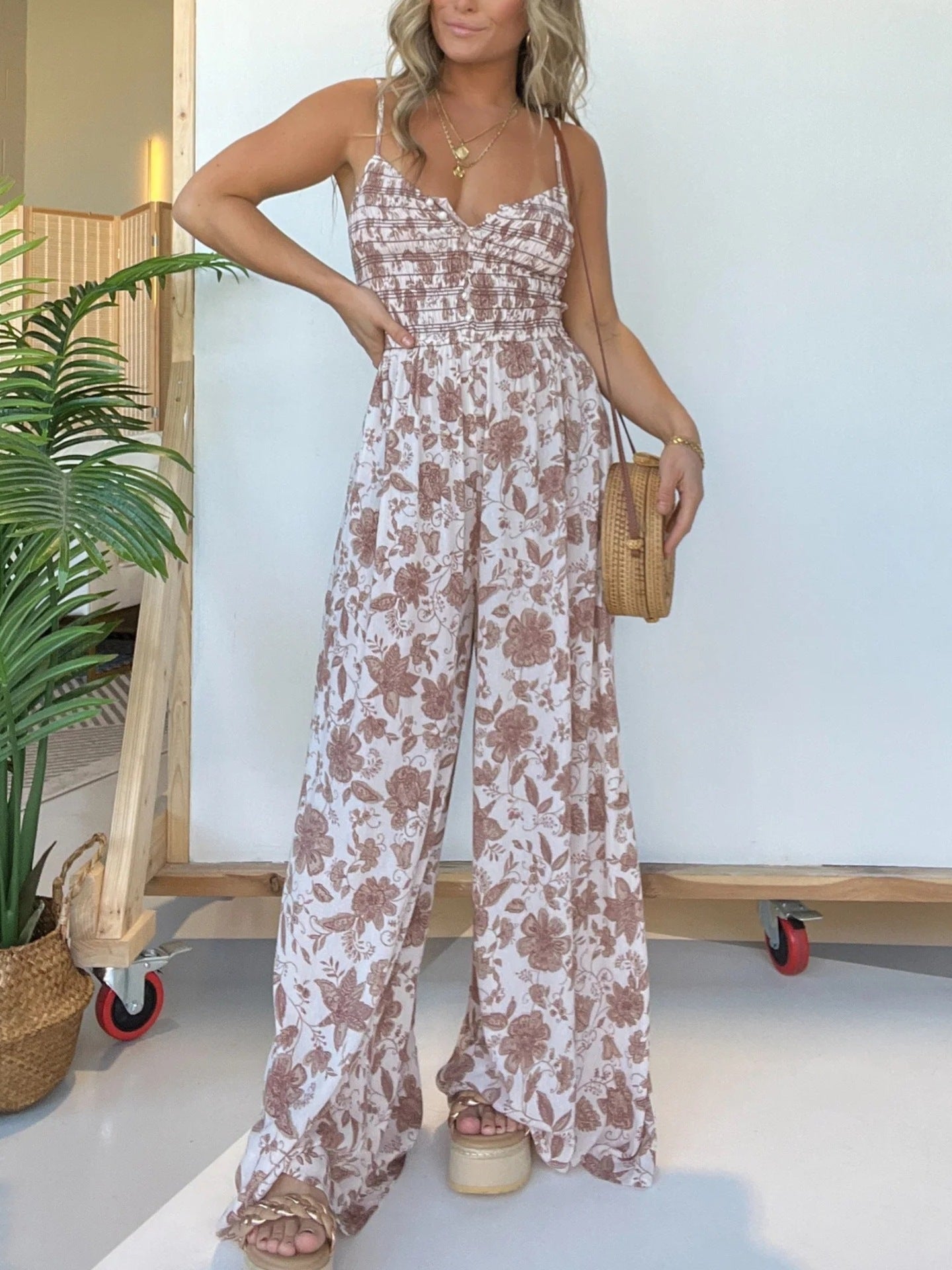 Woman wearing a floral jumpsuit standing indoors with a plant and wooden cart in the background.