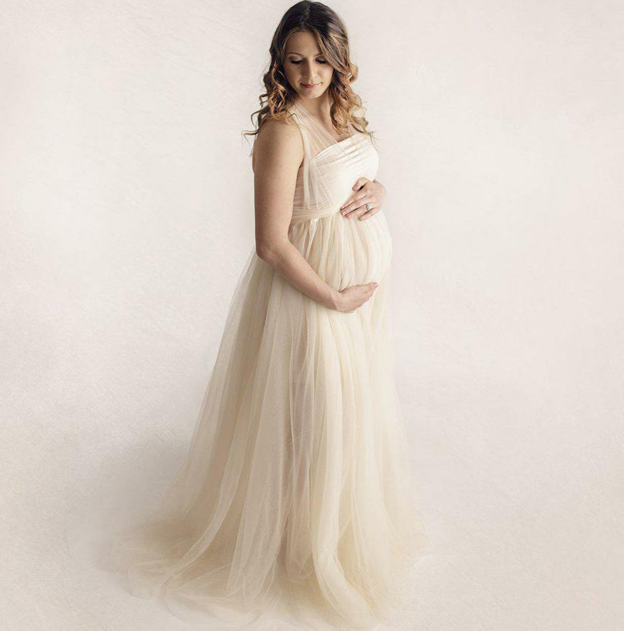 Woman in a flowing white dress holding her pregnant belly against a plain background