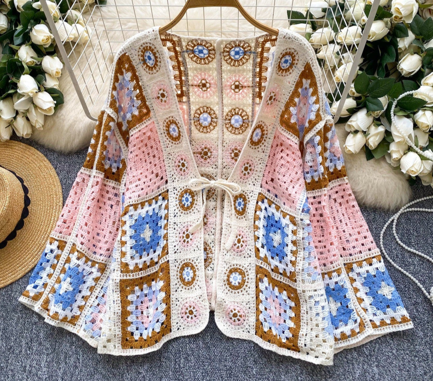 Crocheted cardigan with geometric pattern on a hanger surrounded by flowers and a hat.