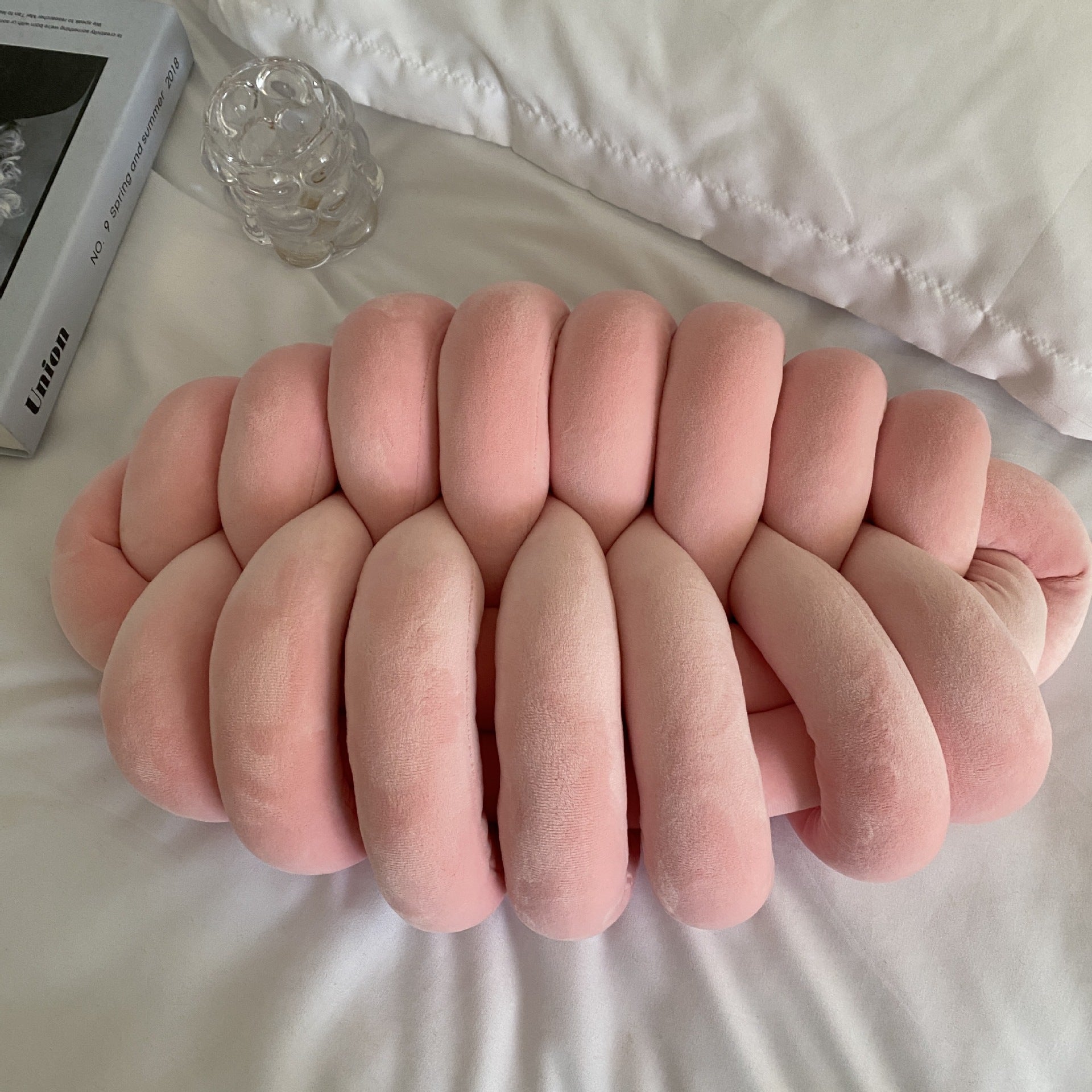 Pink knitted pillow on a white surface with a book and glass in the background