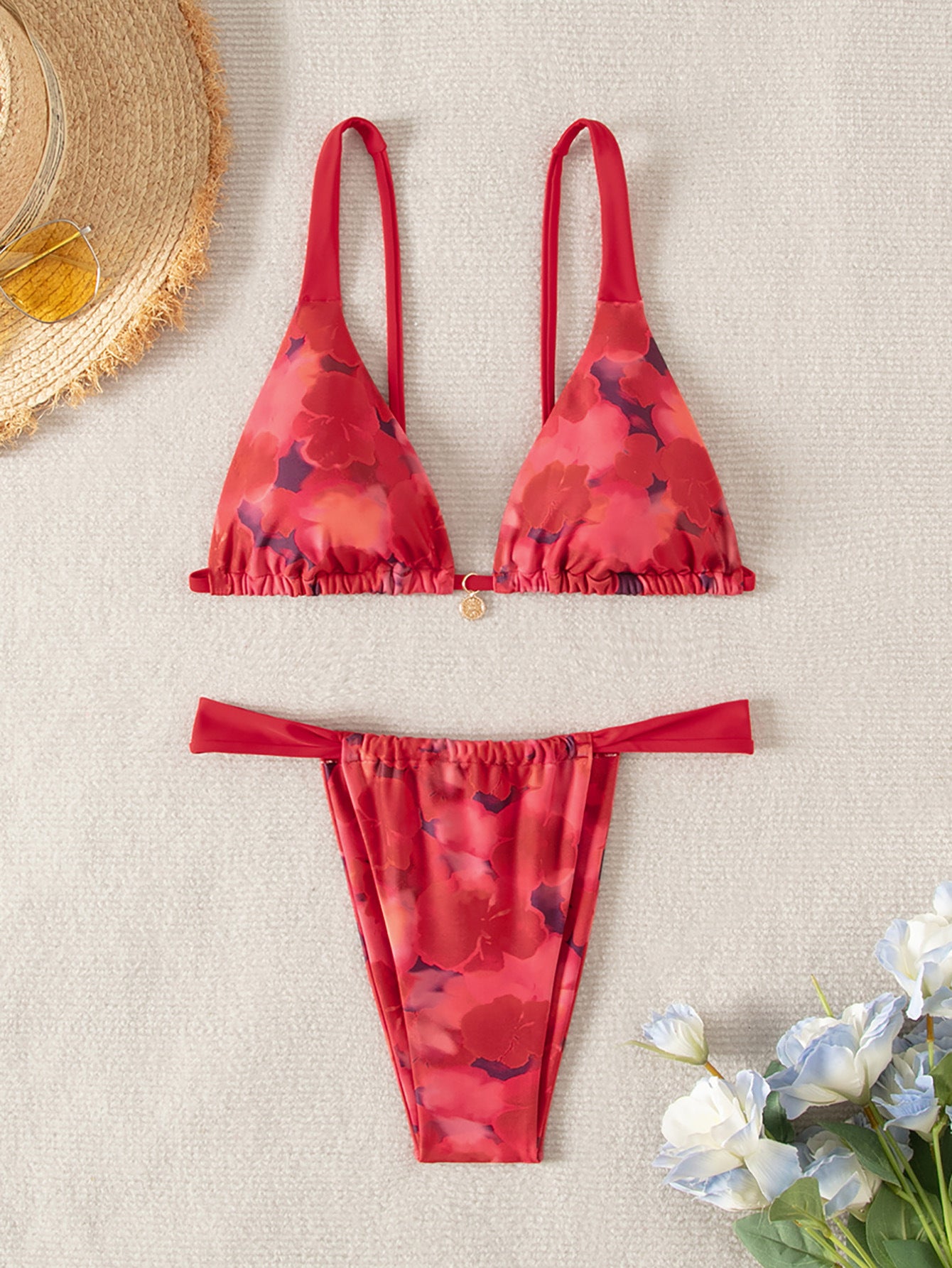 Red floral bikini set on a beige surface with a straw hat and flowers.
