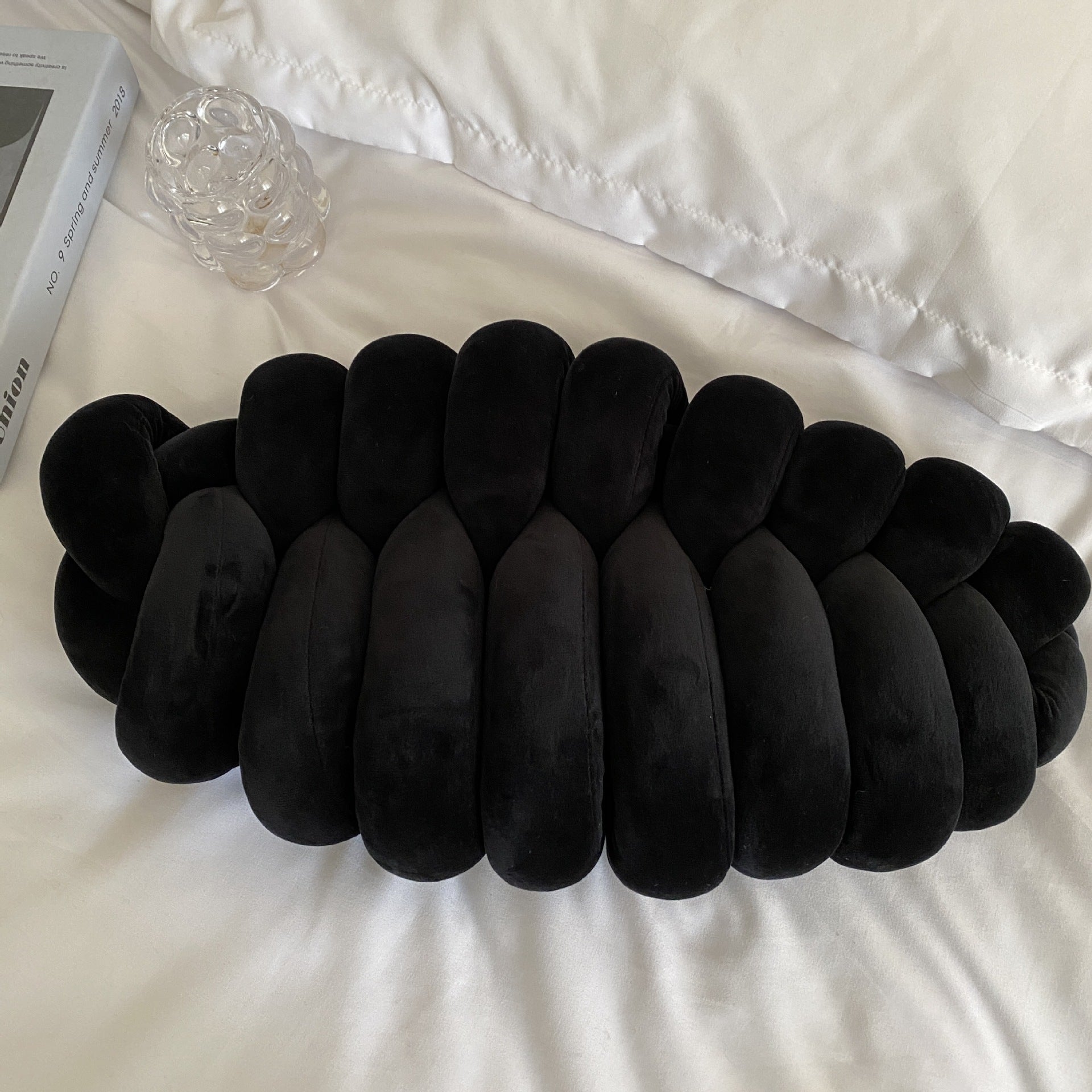black textured pillow on a white surface with a book and glass object in the background