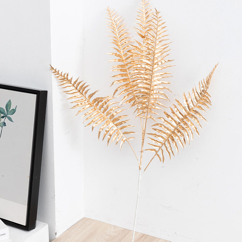 Gold fern plant against a white wall with a framed picture on the left.