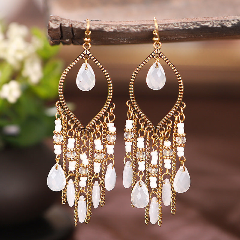 Gold and white teardrop earrings hanging on a wooden stick with a blurred natural background.