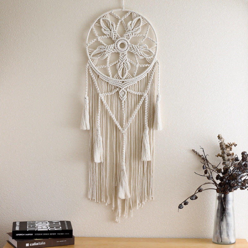 Macrame wall hanging with tassels on a plain wall, next to a vase with dried flowers and books.